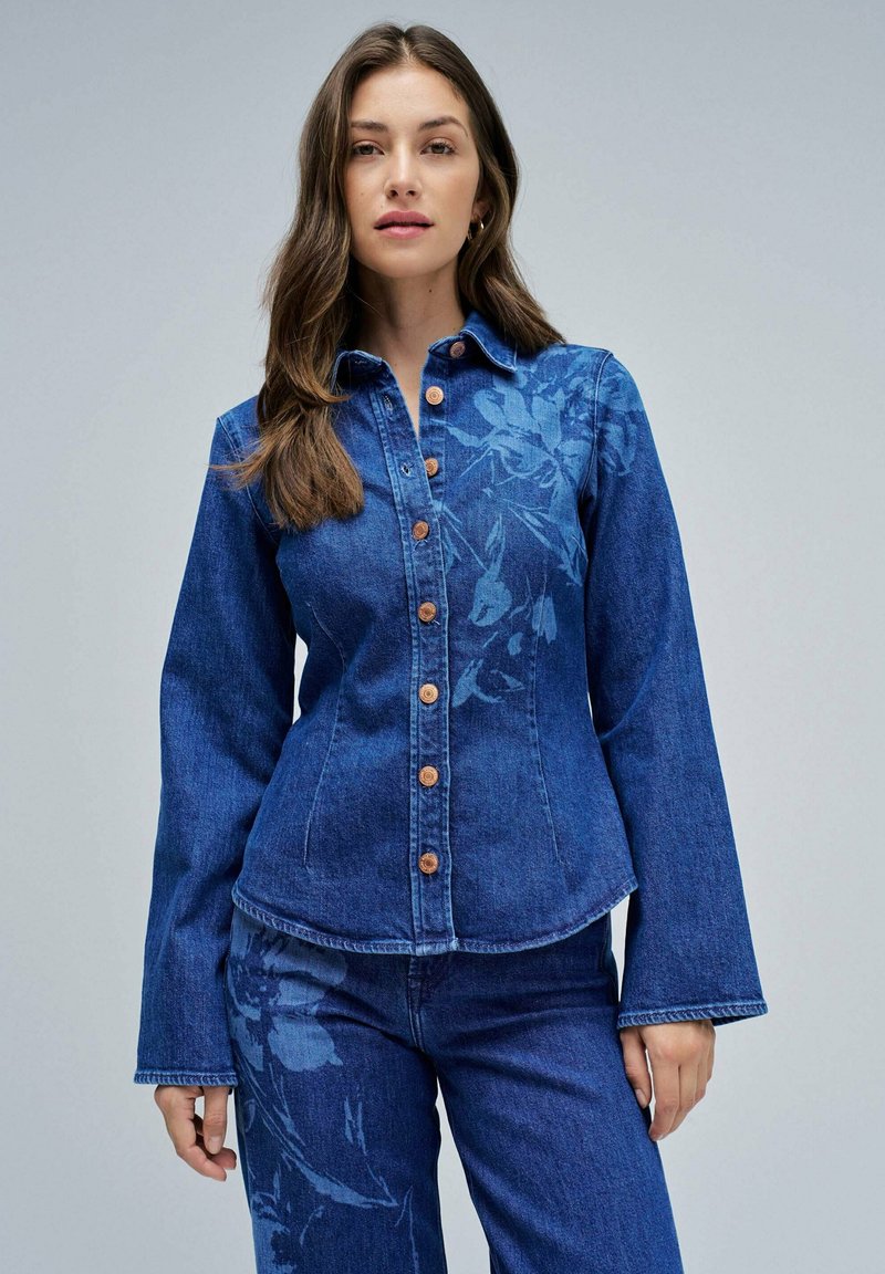 Veste en denim ornée d'un motif floral dans un bleu clair, avec une coupe ajustée, une fermeture à boutons et des manches évasées. Des boutons en ton cuivré accentuent l'avant.