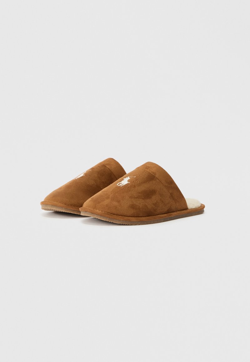 Pantofole in suede marrone con retro chiuso, foderate in morbido beige e con un logo ricamato bianco su un lato. Profilo basso e texture morbida.