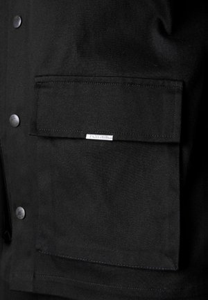 Black jacket with a denim texture, featuring a large front pocket and metal snap buttons. Subtle brand label stitched on the pocket.