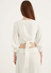 Top bianco avvolgente con maniche lunghe e gonfiate, polsini elasticizzati e dettaglio legato sulla schiena. Tessuto liscio dal design minimalista.