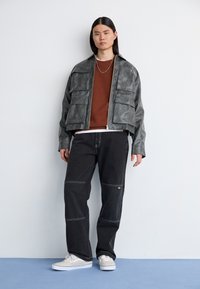 Jachetă gri din piele cu fermoar și buzunare frontale, pulover maro cu gât rotund, pantaloni negri cu lățime mare, cu cusături vizibile, și pantofi casual albi.