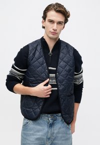 Νέος άνδρας φορώντας ένα ναυτικό γαλάζιο γιλέκο πάνω από ένα μαύρο πουλόβερ με σχέδιο και ανοιχτό μπλε τζιν, στραμμένος μπροστά με ουδέτερη έκφραση.