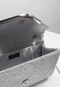 Pochette de soirée en paillettes argentées ouverte pour montrer un intérieur en satin, une chaîne en bandoulière et une étiquette de marque "Mascara London".