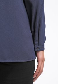 Détail de la manche et de l'ourlet d'une chemise bleu foncé à manches longues avec un poignet à un seul bouton portée par une personne dont la main est détendue le long du corps.