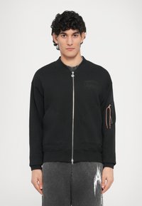 Sudadera negra con cremallera, cuello redondo, dos bolsillos frontales y un bolsillo con cremallera adicional en la manga con cordón de contraste.