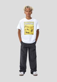 T-shirt bianco a maniche corte con una grafica gialla che recita "FREEDOM" sul davanti. Pantaloni grigi scuri a gamba larga e sneakers bianche.