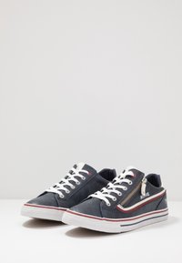 Baskets en toile bleu marine avec lacets blancs, accents rouges et un détail de fermeture éclair latérale. Semelle en caoutchouc avec un motif texturé pour l'adhérence.