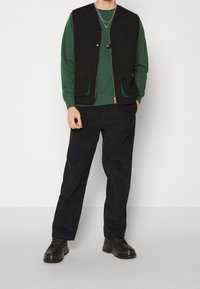 Schwarze ärmellose Weste über einem grünen Pullover, kombiniert mit dunklen Hosen und schwarzen Schuhen. Die Weste hat vordere Taschen und einen goldenen Reißverschluss.