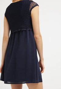Robe bleu marine avec un corsage en dentelle crochetée et des manches courtes, jupe fluide avec une texture lisse, se termine au-dessus du genou.
