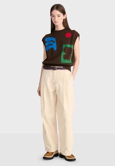 Jeune femme portant un haut marron sans manches avec des formes abstraites, un pantalon beige à jambes larges, une ceinture à motifs et des chaussures beige devant un fond uni.