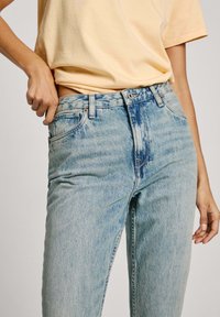 Gaiši zili augstā vidukļa džinsi ar izbalējušu tekstūru, pieci kabatas un oranžas dūriena detaļas; kombinēti ar gaiši dzeltenu T-kreklu.