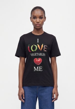 Femme portant un t-shirt noir avec le texte "I love vegetables" utilisant des légumes et un graphique de cœur en tomate, associé à un jean bleu.