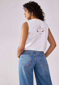Top senza maniche in cotone bianco con bordi scallop e un design a fiori ritagliato sul retro, abbinato a jeans in denim blu a vita alta.