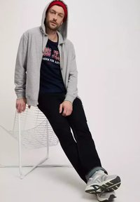 Szary bluza z zamkiem, granatowy T-shirt z napisem, czarne spodnie, szare trampki. Model siedzi na białym, drucianym krześle, neutralne tło.