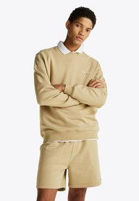Sudadera beige con cuello redondo y logo bordado, acompañada de pantalones cortos a juego. Tejido suave con una textura lisa; ajuste casual.