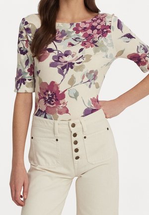 Kobieta ubrana w dopasowaną bluzkę w kwiatowy wzór z fioletowymi i różowymi kwiatami, zestawioną z kremowymi spodniami z wysokim stanem zapinanymi na pięć guzików.