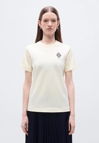 Ανοιχτόκίτρινο βαμβακερό μπλουζάκι με στρογγυλή λαιμόκοψη, κοντά μανίκια και μαύρο λογότυπο σε σχήμα διαμαντιού στο αριστερό στήθος.