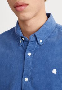 Camisa azul de pana con cuello abotonado, botones blancos y un pequeño logo blanco. La tela tiene un patrón texturizado y acanalado.