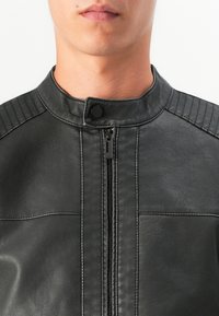Homme portant une veste en cuir noir avec une fermeture éclair à l'avant et un col à bouton-pression, visible du menton au torse.