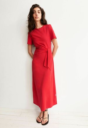 Femme aux cheveux longs et bouclés portant une robe midi rouge à manches courtes avec un lien sur le côté, debout contre un mur blanc et chaussée de sandales noires.