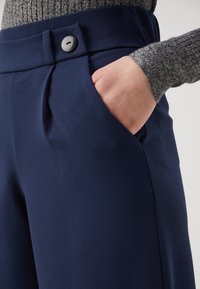 Námořnické modré kalhoty s hladkým materiálem, předním záhybem a šedým knoflíkem. Ruka v kapse, ukazuje strukturu a design kalhot.