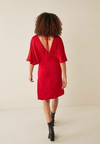 Robe rouge à manches courtes avec un dos en V profond et des détails froncés à la taille. Tissu lisse et coupe décontractée. Bottines noires complètent le look.