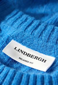 Niebieski sweter z dzianiny z prążkowaną fakturą i metką z napisem "Lindbergh RELAXED FIT" wyboldowanym czarnym fontem na białym tle.