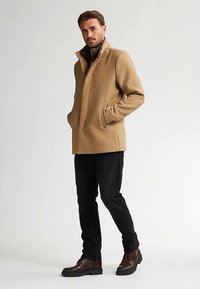 Beige villakangastakki, jossa korkea kaulus ja sivutaskut, päällä tumma pooloneule, yhdistetty mustiin housuihin ja ruskeisiin nahkasaappaisiin.