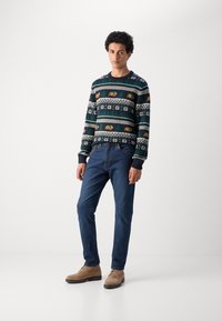 Gebreide trui in donkerblauw met een patroon in rood, wit en groen, gecombineerd met blauwe spijkerbroek en beige schoenen.