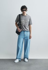 T-shirt cinzenta de algodão com logótipo impresso, combinada com jeans azul claro de pernas largas. Acessórios incluem uma bolsa preta de tiracolo e ténis brancos.