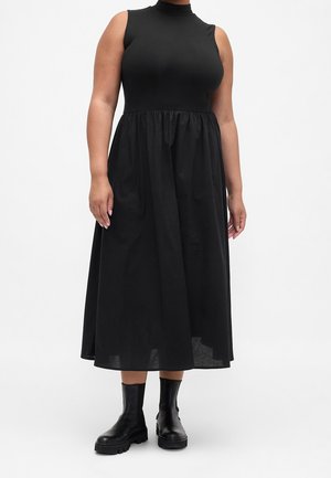 Vrouw draagt een mouwloze zwarte midi-jurk met hoge hals en geplooide taille en zwarte enkellaarzen, staande op een witte achtergrond.