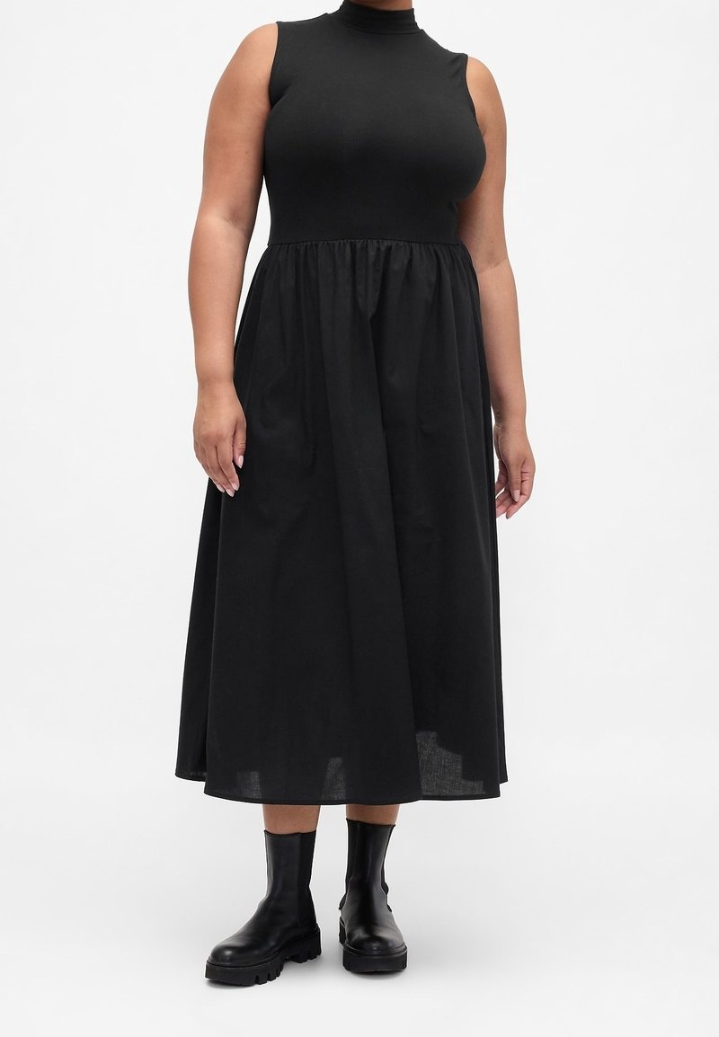 Femme portant une robe midi noire sans manches à col montant avec taille froncée et bottines noires, debout sur fond blanc.