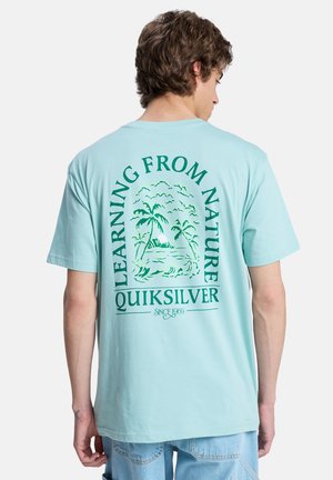 Lyseblå bomulds-T-shirt med grønt grafiktryk af palmer og teksten "Learning From Nature" på bagsiden.