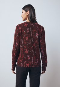 Pull marron avec un motif festif de nœuds rouges, bouteilles et dessins ludiques. Manches longues et texture en maille côtelée.