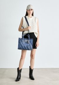 Denim schoudertas met twee handvatten aan de bovenkant, blauw verlooppatroon, gestikte logo "Coach New York," vergezeld door een persoon die een gebreide top en een zwarte rok draagt.