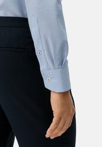 Světle modrá košile s texturovaným vzorem, zapnutými manžetami na knoflíky a tmavě modré kalhoty. Tkanina vypadá hladce a střih je na míru.