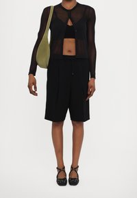 Černý průsvitný cardigan s dlouhým rukávem přes černý bralette, doplněný černými plisovanými šortkami. Doplňky tvoří zelená kabelka přes rameno a černé boty.