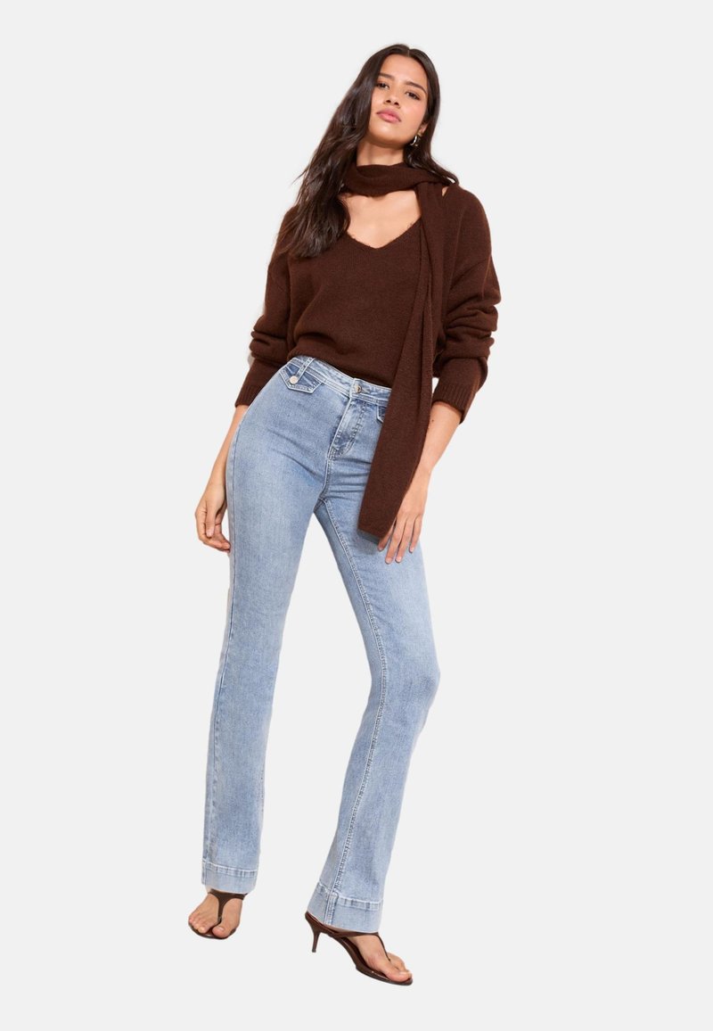 Kvinde iført en brun sweater, matchende tørklæde, lyseblå højtsiddende jeans og brune åbne hæle-sandaler, stående mod en ensfarvet baggrund.