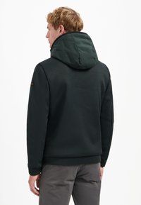 Man in een donkere gestructureerde jas met een gewatteerde capuchon, staande met zijn rug naar de kijker op een effen witte achtergrond.