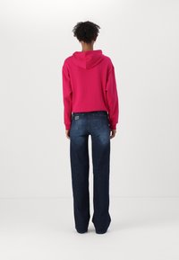 Osoba stojící zády, oblečená v jasně růžové mikině s kapucí a tmavě modrých rovně střižených džínách, na jednobarevném bílém pozadí.