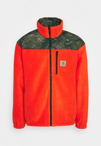 Oranžová fleecová bunda s horní částí se vzorem maskování, vysokým límcem, černým zipem a bočními kapsami. Na hrudi je logo.