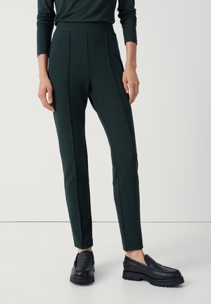 Pantalons ajustés vert foncé à taille haute, avec de vives plis et une texture lisse, associés à des mocassins noirs épais.