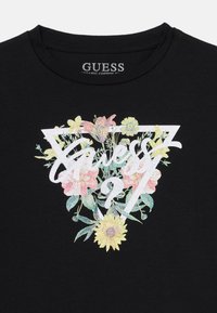 Svart bomull T-skjorte med blomsterdesign som viser pastellfarger og teksten "Guess 27" i holografisk hvit innenfor en triangelramme.