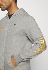 Sudadera con cremallera color gris que presenta una textura suave, capucha ajustable y letras doradas "LAKERS" en la manga derecha. Logo pequeño en negro en el pecho.