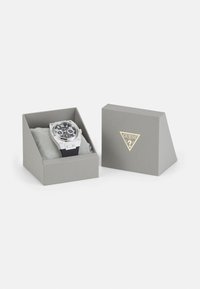 Reloj negro con acentos plateados en una caja gris. Las características incluyen números grandes, un bisel texturizado y una correa de silicona negra.