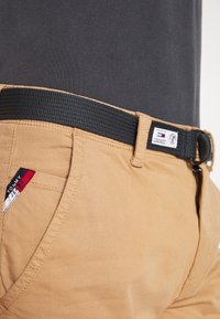 Khaki katoenen broek met een zwarte gestructureerde riem, ritssluiting en een rood-blauwe logo-label aan de zijnaad.
