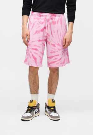 Férfi rózsaszín batikolt rövidnadrágban, fekete hosszú ujjú ingben, fehér zokniban és többszínű magas szárú tornacipőkben egy egyszínű háttér előtt áll.