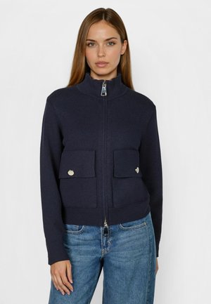 Frau trägt eine marineblaue Strickjacke mit Reißverschluss, Klappentaschen und silbernen Knöpfen, kombiniert mit blauen Denim-Jeans, steht vor einfachem Hintergrund.