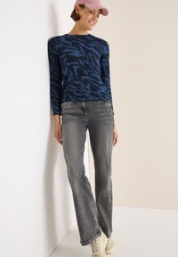 Blau und schwarz gestreifter Langarmpullover kombiniert mit grauen Jeans und hellen Sneakers, vor einem neutralen Hintergrund.