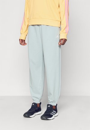 Personne portant un pantalon de survêtement Adidas bleu clair, des baskets Adidas noires et un sweat jaune avec des rayures roses sur les manches, debout devant un fond uni.